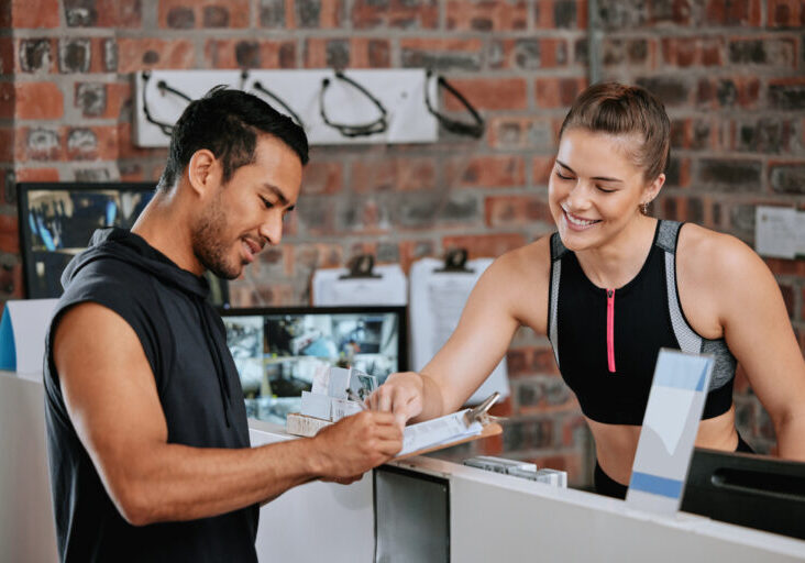 Asian man signing up for gym membership with caucasian trainer. Young coach behind reception pointi.